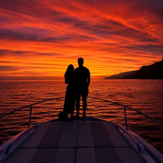 A couple standing on the bow of the boat against a deep red Madeira sunset